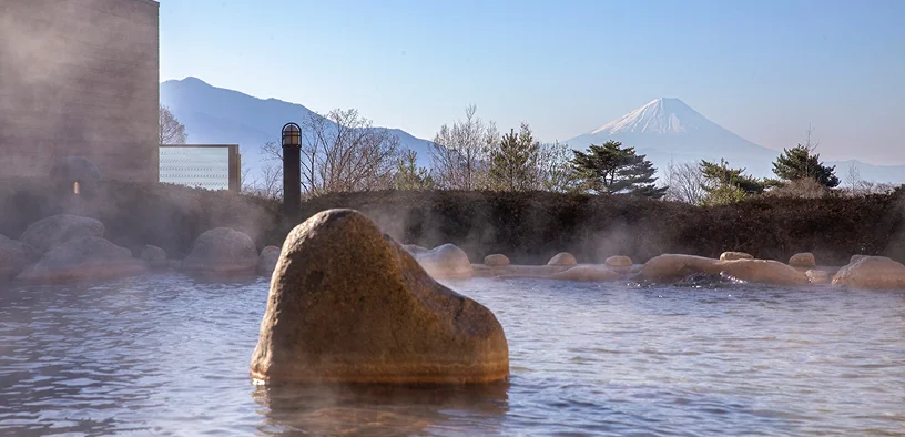 露天風呂越しに大きな岩と湯けむり、背景に木々と晴れた空、右奥に富士山がくっきりと見える風景（パノラマの湯）